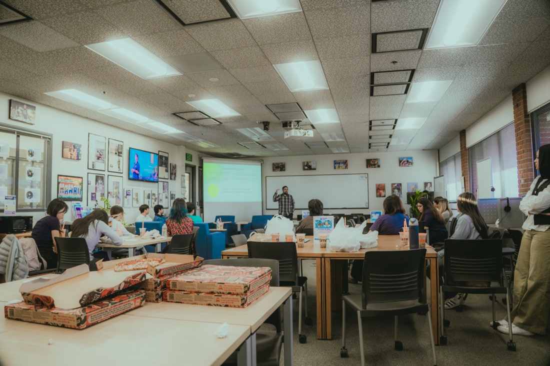 A classroom of fellows at tables with pizza boxes and drinks, watching a presenter at the front