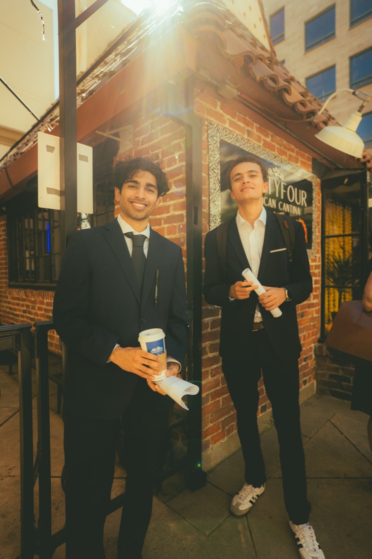 Two fellows in suits holding coffee and rolled papers outside a brick restaurant storefront